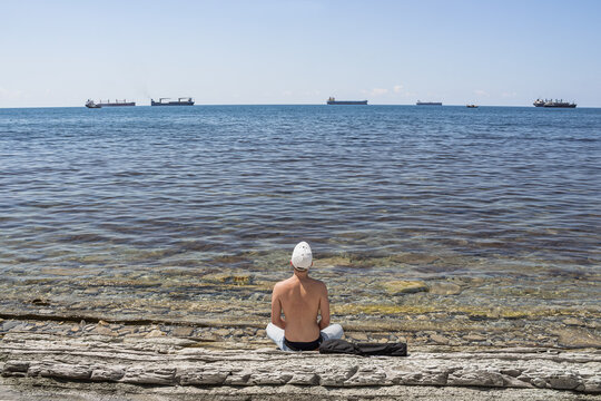 Summer sea landscape. A guy sits on a stone wild beach and looks into the distance. On the horizon is a clear blue sky and cargo ships. The resort city of Gelendzhik. Russia, Black Sea coast