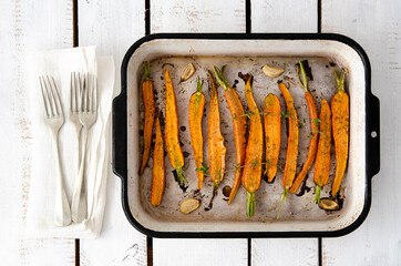 Roasted carrots with garlic, oil and thyme, in a rectangular tray with three forks and napkin, on a white wooden backdrop. Top view. 