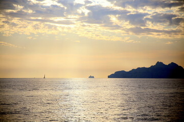 Sunset light on an isolated sailboat, sailing on the horizon, with the coast in backlight, under a dramatic sky