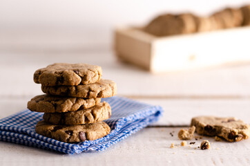 Vanilla and chocolate cookies stacked on a white and blue napkin, a bitten cookie and cookies in a wooden box, on a white wooden backdrop.