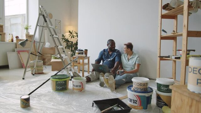 Zoom in shot of multiethnic married couple sitting together on floor in room under renovation, using laptop and discussing remodeling ideas