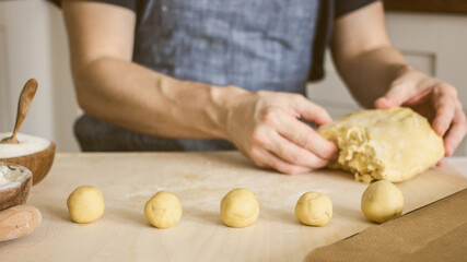 A man without a face in an apron makes homemade shortbread dough cookies on the kitchen table and rolls balls. Around ingredients for baking. Concept of authentic home hobby baker.