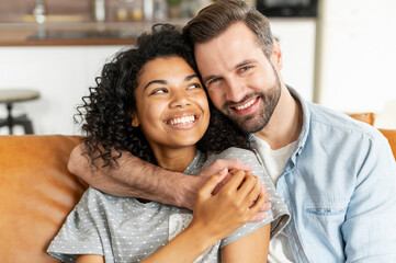 Hilarious multiracial couple in love sits in embraces in cozy living room at home. A close-up portrait of smiling caucasian guy and charming african girl are hugging. Love and affection concept
