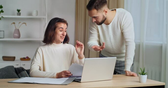 Young woman wife sits at home table looks at laptop has problems with paperwork makes mistake in document serious man husband approaches argues indignant reads letter feels anger on silly girl