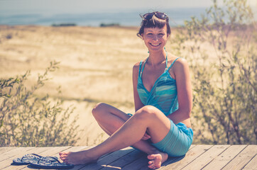 Young beautiful girl in bikini, pareo wrap and sunglasses on head sitting on wooden path, smile and...