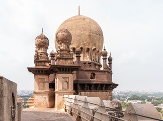 Vijayapura, Karnataka, India - November 8, 2013: Gol Gambaz Mausoleum. Horizontal view on beige stone top of one tower under light blue cloudscape. Cityscape down there. © Klodien