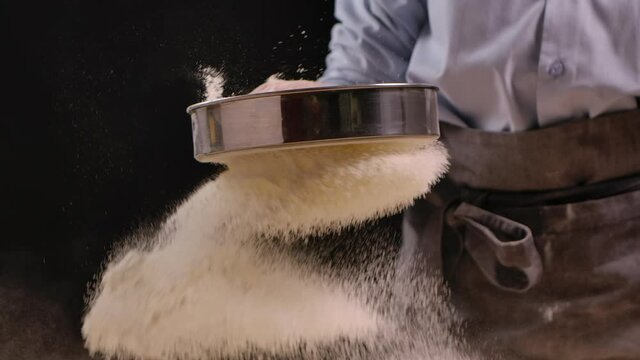 Woman is sifting flour through sieve for baking. White powder flying in different directions on a black background. Preparing flour to baking cake in kitchen at home. Close up. Slow motion.