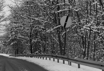 Black and white photo of "End of speed limit" road sign next to forest road in snowfall winter scenery