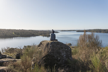 Young and attractive female meditating on top of a rock and in front of a lake during a sunny and cold winter morning