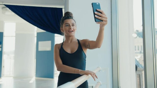 Young beautiful woman standing in gym near ballet machine and holds smartphone talking on video call with friends.