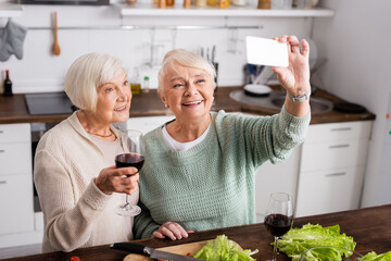 cheerful senior woman holding glass with red wine and looking at smartphone while friend taking selfie in kitchen