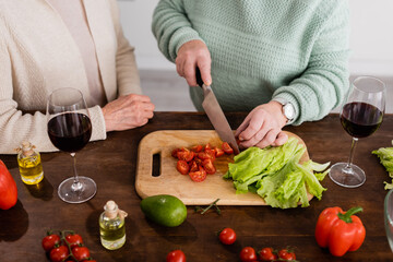 cropped view of retired woman cutting cherry tomatoes on chopping board near senior friend