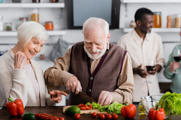 happy senior woman pointing with finger at retired man cooking salad near multicultural friends on blurred background