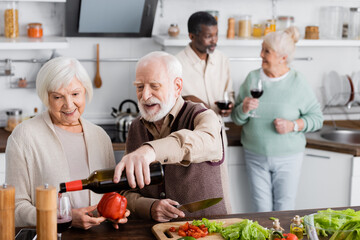 happy senior man pouring wine in glass near retired woman and multicultural friends on blurred background