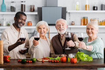 joyful multicultural pensioners holding glasses on wine near fresh vegetables on table