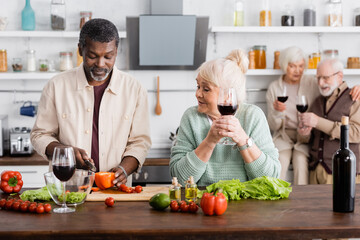 African american man cutting bell pepper near senior woman and retired friends on blurred background