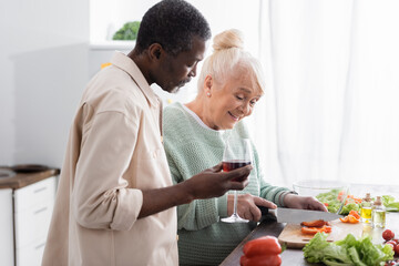 Obraz na płótnie Canvas smiling senior woman cutting bell pepper near african american husband with glass of wine