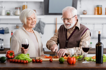 happy senior man cutting vegetables near smiling wife with glass of wine