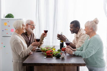 senior interracial friends holding glasses with wine and smiling near vegetables on table