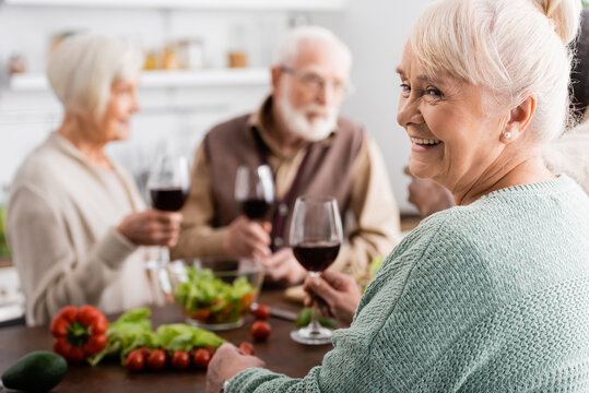 happy senior woman holding glass of red wine near retired friends on blurred background
