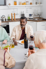 senior african american man holding glass of red wine near retired friends on blurred foreground