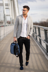 Happy young businessman standing on the bridge. Yuppie outdoor holding bag. Smart young professional walking outside