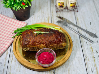 Meat baked on a wooden plate. Meat sauce in a saucepan. Fork and knife in the background. Barbecue meat with a sprig of rosemary.