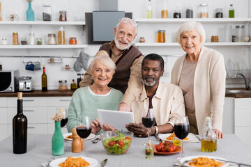 senior multicultural people holding digital tablet near happy friends and tasty food on table