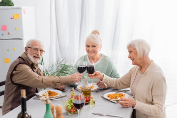happy senior friends clinking glasses with red wine near tasty lunch on table
