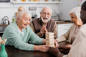 amazed retired woman playing tower wood blocks game near cheerful multicultural friends