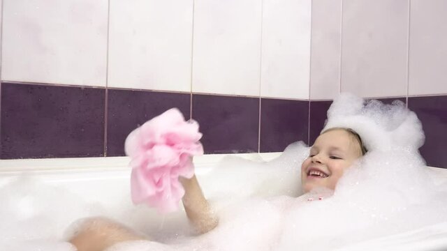 A little girl in a bubble bath lathers her leg with a pink washcloth. Lots of foam.Close-up.