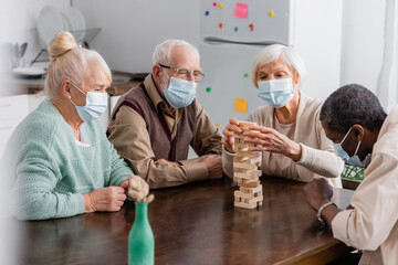 retired interracial friends in medical masks playing tower wood blocks game at home
