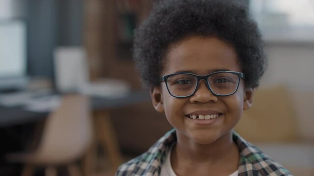 Slow-motion close-up portrait of cute afro kid in glasses smiling to camera with modern living room in blurred background