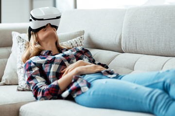 Obraz premium Beautiful young woman using virtual reality headset and enjoying the moment lying on couch at home.