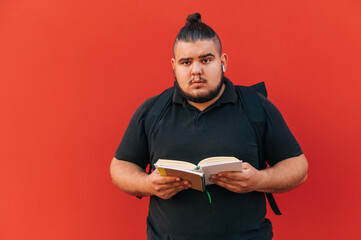 Funny overweight student with a backpack on his back stands with a book in his hands on a red wall background and looks at the camera with a serious face.