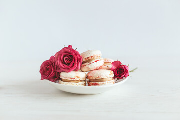 Tasty french macaroons with pink roses on a white plate.  White wooden background.