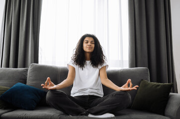 Obraz premium Smiling multiracial black curly girl uses cell phone, talking with friends or family while sits on the couch at living room and looks at camera, communication concept