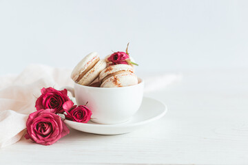 Tasty french macaroons with pink roses in a white cup. White wooden background.