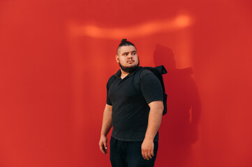 Handsome fat man with a backpack stands on a background of a red wall and looks to the side,...