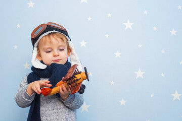 Fototapeta na wymiar Little boy in helmet and glasses with wooden plane against the background of a blue wall with stars. Happy child playing with toy airplane