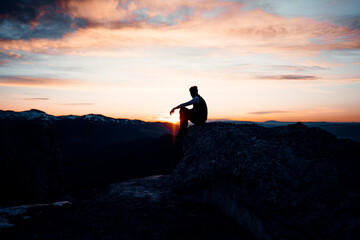 PERSON SITTING AT THE TOP OF A MOUNTAIN. MAN ENJOYING AND HAVING FUN DURING SUNSET. OUTDOOR AND SUNSET CONCEPT.