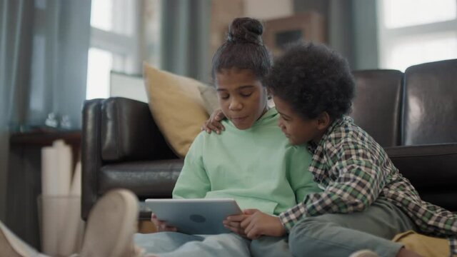 Slow-motion medium close up of joyful afro siblings spending time together playing games on digital tablet sitting on couch in living room