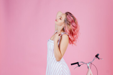 Fototapeta na wymiar Adorable woman in stylish dress expressing happiness during photoshoot near bicycle. Cheerful girl with pink hair laughing while posing on pink background.