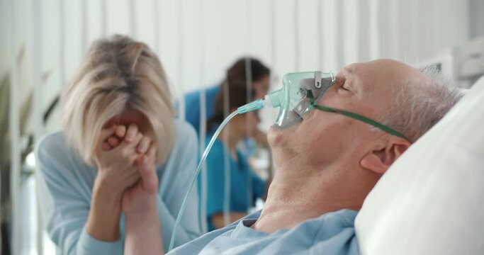 Crying woman holding hand of aged husband patient while sitting by bed at hospital