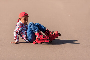 Happy little african american boy on rollers skates at asphalt road. Roller kid fell and smiles sitting on ground and trying to get up. Risk of injury not wearing protection.