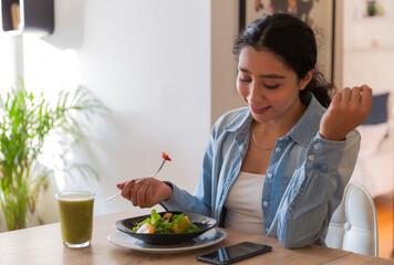 Young brunette woman eating a salad in her dinning room. She is using her phone