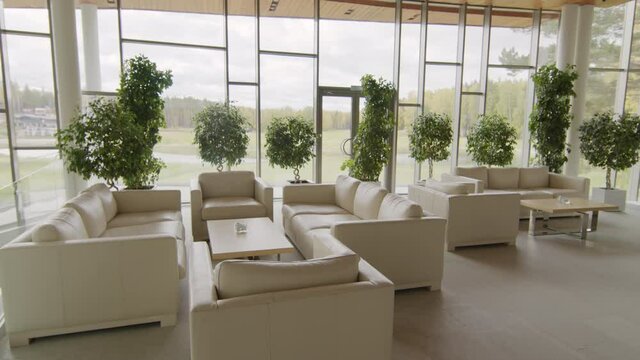 No people arc shot of posh lounge zone with beige leather sofas and panoramic windows with picturesque view on green lawn