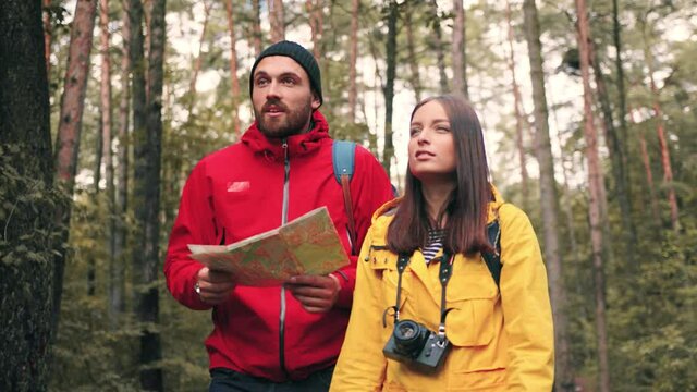 Young stylish couple of tourists walking in park studying paper map looking around and talking. Concept of hiking and outdoor activities.