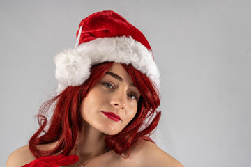 Young female attractive model with auburn hair and red lipstick sat down in a studio with a white background wearing a sexy santa outfit with a red Santa hat with red velvet gloves.