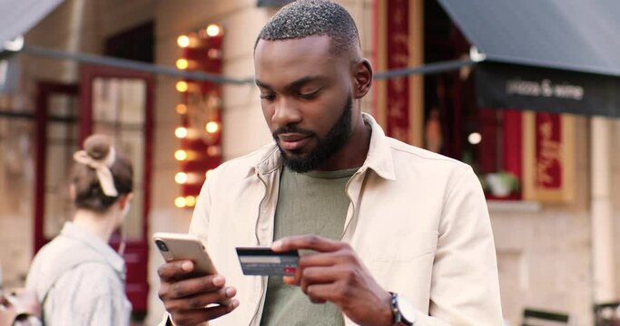 Young African American stylish man standing at street and tapping on smartphone while shopping online with credit card. Handsome male buying and paying in internet on mobile phone. Outside.